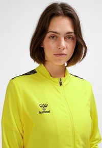 Helles gelbes Reißverschlussjacke aus strukturiertem Stoff, mit schwarzen Musterakzenten auf den Schultern und einem Logo auf der linken Brust.