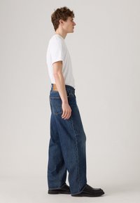 Jonge man die zijwaarts staat, gekleed in een wit T-shirt met korte mouwen, losse donkerblauwe jeans en zwarte instappers, met een effen achtergrond.