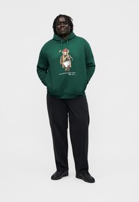Moški s temno poltjo in kratkimi prameni dreadlocks, ki nosi zelen pulover s kapuco z grafiko medveda, črne hlače in črne čevlje, stoji z rokami v žepih.