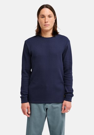 Personne debout portant une chemise texturée bleu marine à manches longues et un pantalon bleu clair sur fond blanc uni.