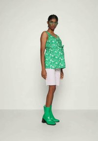 Top sem mangas com padrão floral verde e design franzido, com um laço na cintura, combinado com calções roxos claros e botas de tornozelo verdes brilhantes.