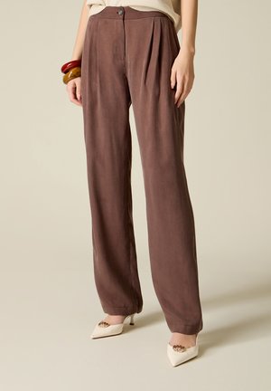 Donna che indossa pantaloni marroni a vita alta e gamba larga, tacchi crema con decorazioni dorate, e braccialetti spessi rossi e color senape.