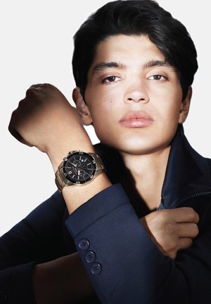 Montre noire avec accents en argent, bracelet en acier inoxydable et cadran superposé sur un poignet. Le modèle porte une veste marine avec des détails de boutons.