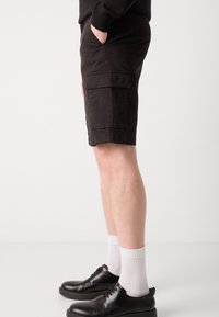 Svarte cargo shorts med flate lommer, laget av bomull, knelange, kombinert med svarte sko og hvite sokker, vist fra en sidevinkel.