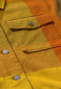 Tela a cuadros amarilla y naranja con un acabado texturizado; presenta un bolsillo en el pecho con un botón de presión plateado y detalles de costura visibles.