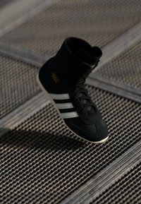 Zapatilla alta negra con rayas blancas y logo dorado, con un upper texturizado, cordones y una suela de goma en contraste.