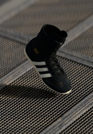 Sneaker alta nera con strisce bianche e logo dorato, caratterizzata da una tomaia testurizzata, lacci e una suola in gomma a contrasto.