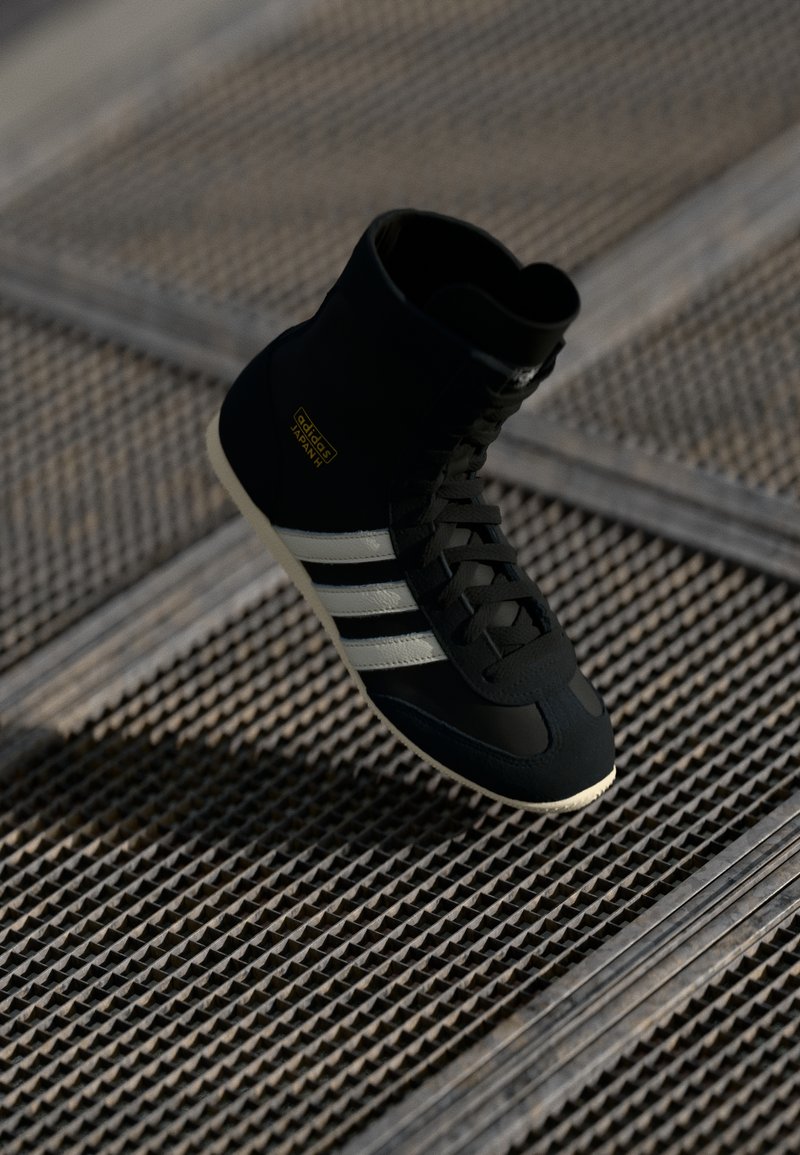 Zapatilla alta negra con rayas blancas y logo dorado, con un upper texturizado, cordones y una suela de goma en contraste.
