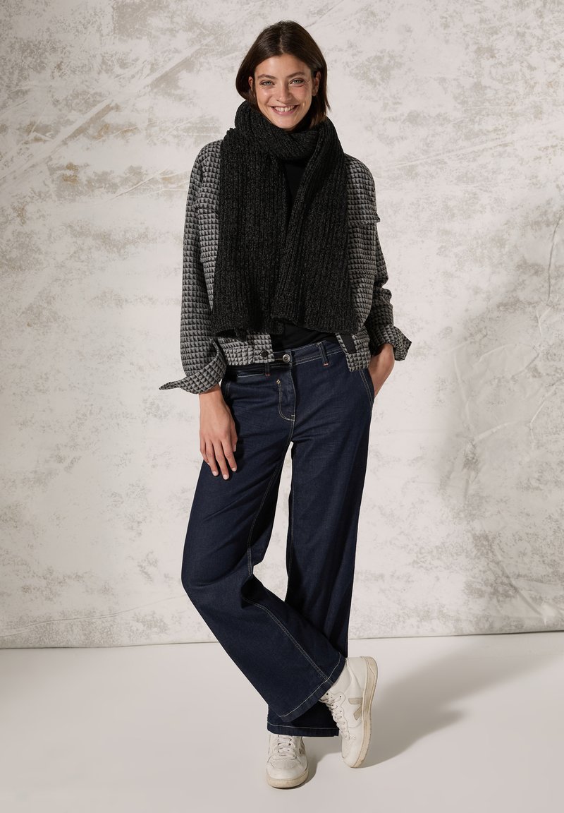 Écharpe noire texturée, chemise à carreaux gris, haut noir, jean large bleu marine, baskets blanches. Le modèle se tient devant un fond neutre.