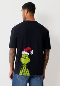 Melna īsroku krekla no kokvilnas ar zaļu Grinča attēlu, aizmugurē ar sarkanu Ziemassvētku vecīša cepuri, brīva piegriezuma.