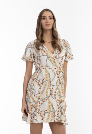 Vrouw met lang golvend haar, gekleed in een witte wikkeljurk met oranje en grijze kettingpatronen en een gouden kettingketting.