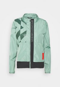 Ljusgrön jacka i glänsande tyg, med ett geometriskt mönster i mörkgrön, framzipper, och svart nederdel med en röd detalj.
