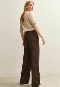 Bruine, gestreepte wijde broek met een elastische tailleband, gecombineerd met een zachte, beige gebreide trui. Model gezien vanaf de achterkant.