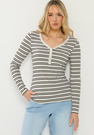 Lyshåret kvinde iført en tætsiddende, langærmet hvid top med sorte horisontale striber og lyseblå jeans, stående med den ene hånd i lommen.