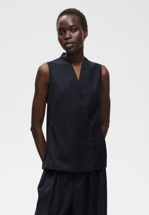 Femme aux cheveux courts portant un gilet noir sans manches à double boutonnage et un pantalon noir assorti, debout les mains dans le dos.