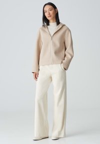 Hellbeige Zip-Hoodie aus weichem Material, kombiniert mit cremefarbenen weit geschnittenen Hosen. Glatte Textur, minimales Design und keine sichtbaren Muster.