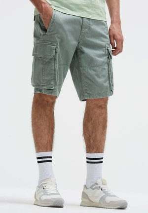 Man wearing light green cargo shorts with flap pockets, white socks with two black stripes, and white-gray sneakers, standing with hands relaxed.