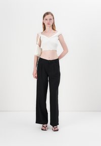 Bílý crop top s volánovými okraji, spárovaný s černými kalhotami s vysokým pasem a širokými nohavicemi; modelka nese malou bílou tašku s tkaným vzorem.