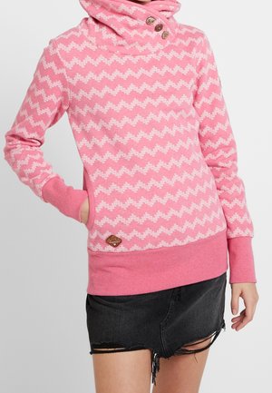 Kobieta nosząca różowy sweter w wzory z guzikami na wysokim kołnierzu oraz czarną, zniszczoną spódnicę jeansową.