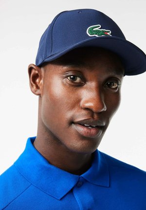 Gorra navy con un logo de cocodrilo verde en la parte frontal, combinada con una camiseta polo azul. Textura suave, forma estructurada y dos botones visibles.