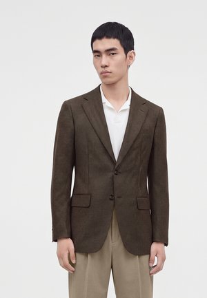 Jeune homme portant un blazer marron à carreaux sur une chemise blanche et un pantalon beige, sur un fond uni.