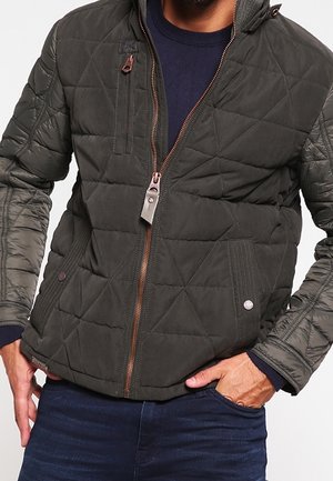 Veste matelassée vert olive foncé avec fermeture éclair à l'avant, poche poitrine et poches latérales à rabat portée sur un pull bleu marine et un jean bleu.