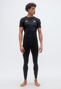 Νέος άνδρας στέκεται ξυπόλητος φορώντας μαύρο εφαρμοστό αθλητικό μπλουζάκι με διακριτικό σχέδιο και μαύρο κολάν που ταιριάζει, κοιτάζοντας προς τα δεξιά.