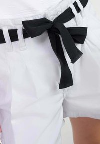 Pantalones cortos blancos con un cinturón de tela negra atado en un lazo, que presentan una textura suave y costuras visibles a lo largo de la cintura.