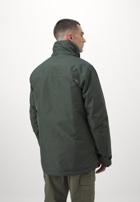Groene waterdichte jas met een hoge kraag, voorzien van een logo op de linkerschouder en een gestructureerde stof, gecombineerd met cargobroek.