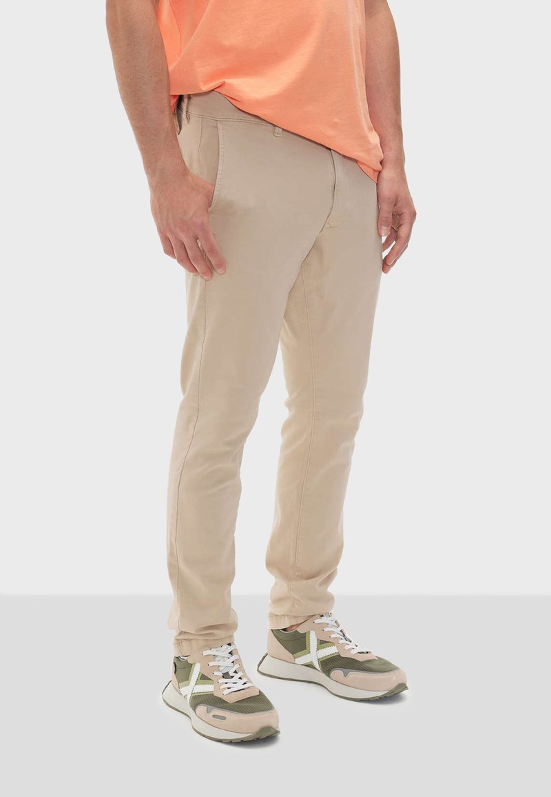 Pantaloni beige slim-fit in cotone, con frontale piatto, due tasche laterali e una texture liscia. Indossati con sneakers verdi e beige.
