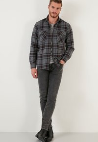 Camicia di flanella a quadri grigia con bottoni frontali, due tasche sul petto, indossata sopra una maglietta grigia, abbinata a jeans scuri e stivali neri.