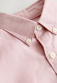 Šviesiai rožinė marškinėlių apykaklė su tekstūruota medžiaga, turinti du baltus mygtukus ir tvarkingas siūles ant apykaklės ir užtrauktuko.