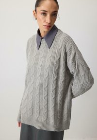 Gai pelēks adīts džemperis ar ribotu apakšmalu un pārspīlētu piegriezumu, zem kura ir redzama kontrastējoša pelēka krekla apkakle.