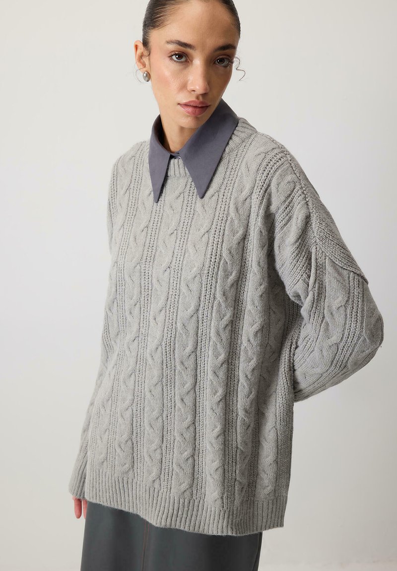 Gai pelēks adīts džemperis ar ribotu apakšmalu un pārspīlētu piegriezumu, zem kura ir redzama kontrastējoša pelēka krekla apkakle.