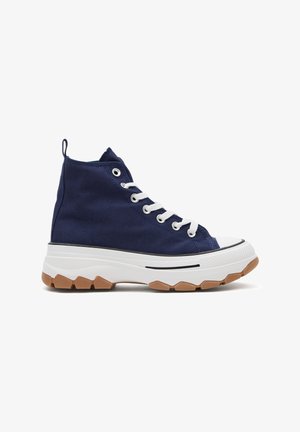 Zapatilla de lona azul marino de caña alta con cordones blancos, suela blanca gruesa y dibujo de goma marrón, mostrada de perfil sobre fondo blanco.
