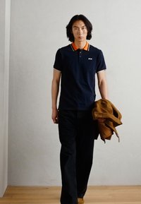 Jūrų jūreivio polo marškiniai su oranžinėmis apykaklės detalėmis, derinami su tamsiomis kelnėmis. Modelis laiko rudą striukę ir vaikšto minimalizmo stiliaus aplinkoje.