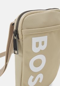 Borsa a tracolla in tessuto beige con grande scritta bianca "BOSS", cerniera nera e tracolla regolabile.