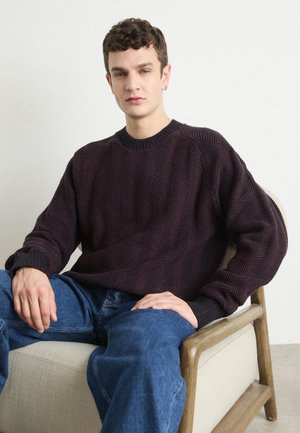 Pulover roșu închis cu model texturat în zig-zag, cu guler și manșete ribbate, purtat cu blugi din denim albastri, așezat pe un scaun deschis.