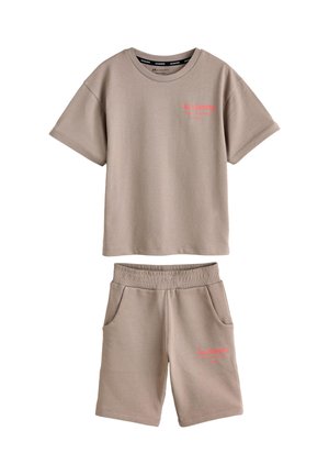 Teenagedreng står med hænderne i lommerne iført beige oversized t-shirt og matchende shorts, hvide sokker og hvide sneakers.