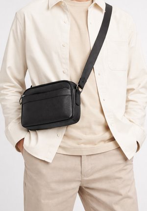 Man wears black leather crossbody bag with adjustable strap over beige shirt and light cream button-up shirt, hands in pants pockets.
