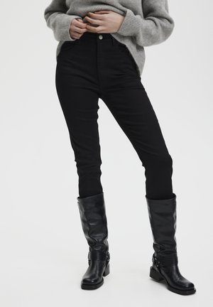 Person iført sorte slim-fit bukser, grå sweater og knæhøje sorte læderstøvler, stående mod en ensfarvet lys baggrund.
