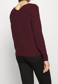 Maglione bordeaux a maglia con scollo a V, tessuto strutturato, maniche lunghe e polsini a costine, abbinato a pantaloni neri, visto da dietro.