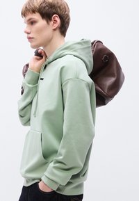 Ανοιχτοπράσινο φούτερ με κουκούλα, τσέπη καγκουρό, κορδόνι κουκούλας και ριμπ μανσέτες. Το μοντέλο κρατά ένα σκούρο καφέ σακίδιο πλάτης στον έναν ώμο.