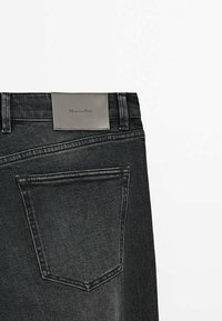 Vue arrière d'un jean en denim noir montrant une seule poche arrière et une étiquette de marque en cuir gris cousue au-dessus de la poche.