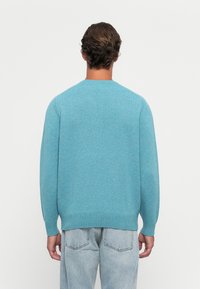 Mann mit kurz gewelltem braunem Haar, der einen blauen Strickpullover und hellblaue Jeans trägt, steht mit dem Rücken zu einem schlichten Hintergrund.