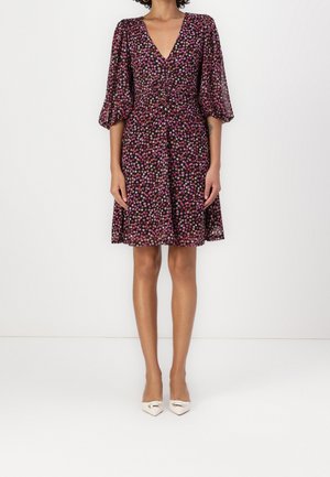 Femme portant une robe noire longueur genoux avec des motifs floraux roses et violets, col en V, taille cintrée et manches trois-quarts bouffantes.