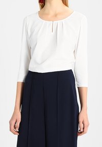 Frau in einer weißen Bluse mit langen Ärmeln und einem kleinen Schlüssel-Ausschnitt vorne, kombiniert mit hochgeschnittenen, marineblauen weiten Hosen.