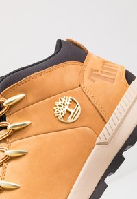 Tenis din suede maro, cu un design înalt, având accesorii de culoare auriu, șireturi roșii și galbene și un logo embosat pe lateral.