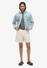 Chaqueta de mezclilla en azul claro, camisa a rayas, pantalones cortos blancos deshilachados, cinturón marrón y sandalias oscuras. Conjunto casual con texturas distintas.