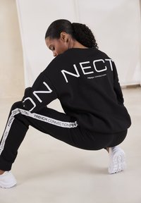 Schwarzer Sweatshirt und Hose mit dem Logo "FRENCH CONNECTION". Der Sweatshirt hat große weiße Buchstaben; die Hose hat weiße Seitenstreifen.
